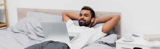 gelukkig Afrikaans amerikaans man in draadloze oortelefoon kijken film terwijl liggend op bed met laptop, banner - Foto, afbeelding