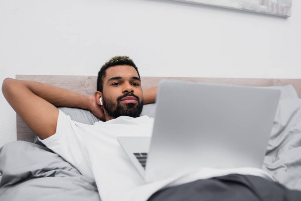 hombre afroamericano en auriculares inalámbricos viendo película mientras está acostado en la cama con el ordenador portátil  - Foto, imagen