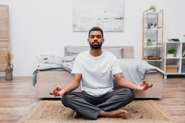 bearded african american man in wireless earphone meditating with closed eyes while sitting on rug near bed - Фото, изображение