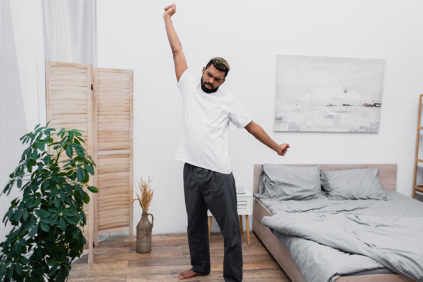 bebaarde Afro-Amerikaanse man stretching in de buurt vouwen scherm in moderne slaapkamer  - Foto, afbeelding