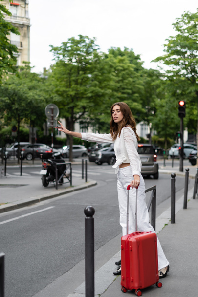 Stylish traveler with suitcase catching taxi near road on urban street in Paris  - Foto, Bild