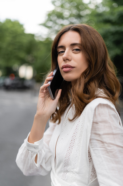 Brunette woman talking on smartphone and looking at camera on street in Paris  - Fotoğraf, Görsel