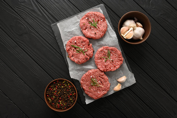 Raw beef burger patties on butcher's wooden board, rosemary and pepper. Black background. View from above. - Foto, afbeelding