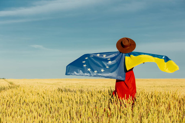 Chica con banderas de Ucrania y la UE en el campo de trigo y el cielo azul en el fondo - Foto, Imagen