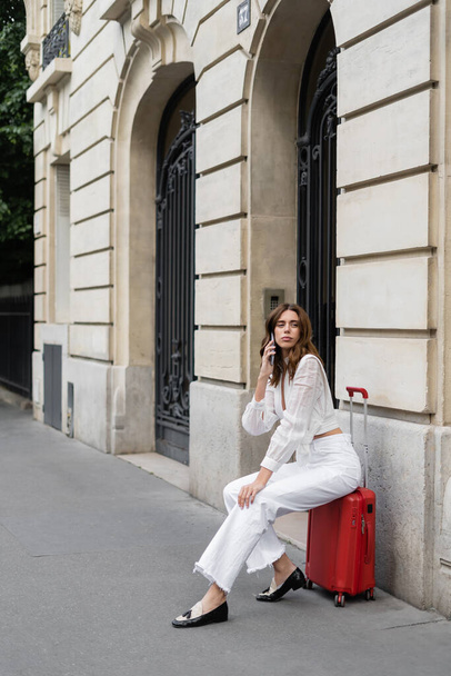 Pretty tourist talking on mobile phone and sitting on suitcase on street in Paris  - Fotografie, Obrázek
