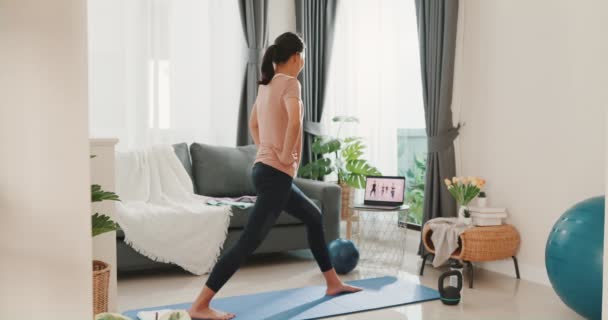 Jonge aantrekkelijke Aziatische vrouw in sportkleding site op mat in de vloer virtuele fitness klasse met een groep mensen op een video conferentie op laptop praktijk yoga voor beginners in de woonkamer thuis. - Video