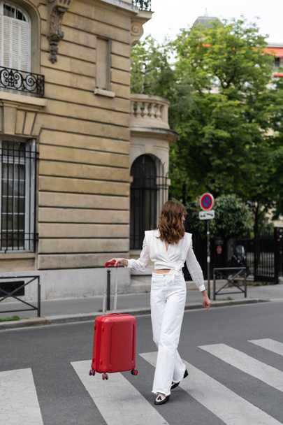 Side view of brunette traveler holding suitcase while walking on crosswalk in Paris  - Foto, afbeelding