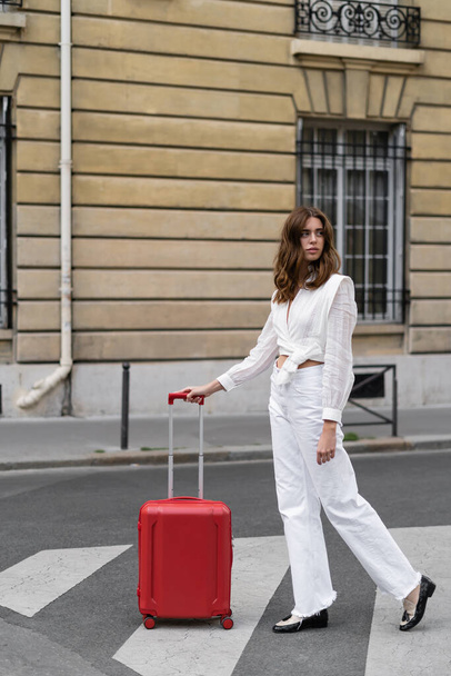 Young woman in blouse standing near suitcase on urban street in France  - 写真・画像