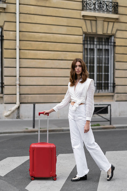 Stylish woman looking at camera near suitcase on crosswalk in Paris  - Fotó, kép
