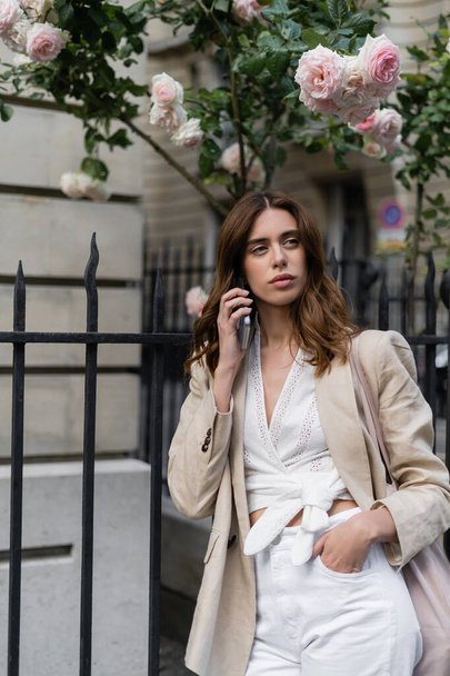 Цікава жінка розмовляє на смартфоні біля квітучих троянд на міській вулиці у Франції.  - Фото, зображення