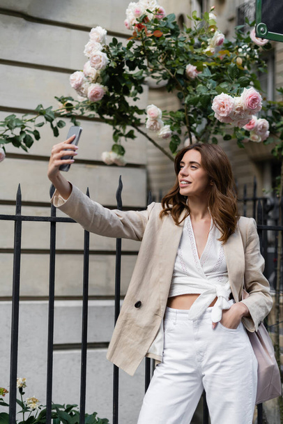 Stylish woman taking selfie on smartphone near blooming roses on street in Paris  - Фото, изображение