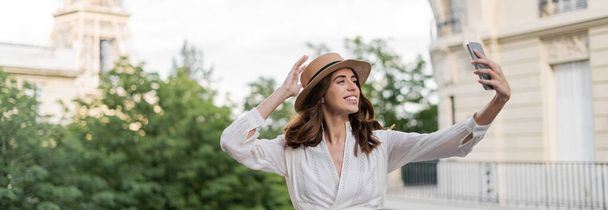 Cheerful young woman in sun hat taking selfie on smartphone with blurred Eiffel tower at background in France, banner  - Fotoğraf, Görsel