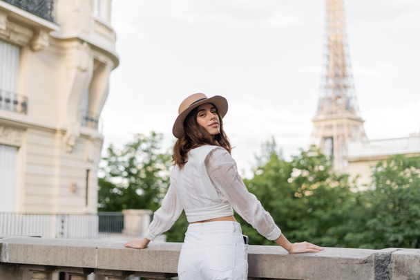 Uśmiechnięta kobieta w kapeluszu przeciwsłonecznym oglądająca kamerę z wieżą Eiffla na tle Paryża  - Zdjęcie, obraz