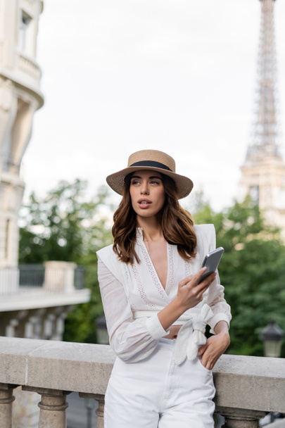 Trendy woman in sun hat holding smartphone with Eiffel tower at background in Paris  - Fotografie, Obrázek