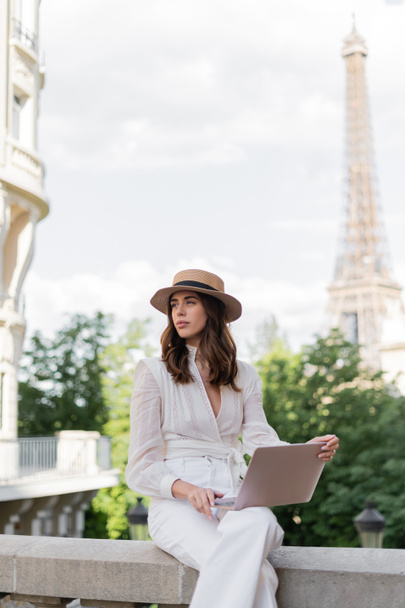 パリを背景にエッフェル塔がぼやけている都市の通りにラップトップを使用して太陽の帽子のファッショナブルなフリーランス  - 写真・画像