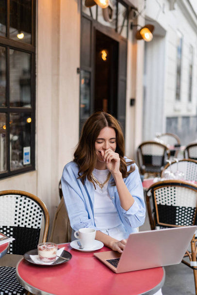 Positive freelancer using laptop near coffee and dessert in cafe on urban street in France  - Фото, изображение