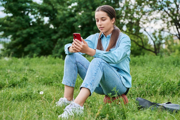 Teenage female student resting sitting on grass using smartphone. Attractive teen girl on lawn with basketball ball, backpack. High school, technology, active healthy lifestyle, adolescence concept - Foto, imagen