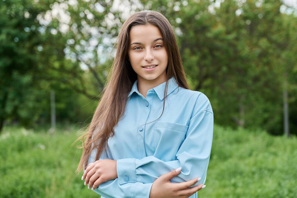 Outdoor portrait of a beautiful teenage girl 15 years old. Smiling female teenager looking at the camera in the park, against the backdrop of green grass. Adolescence, high school, age concept - Φωτογραφία, εικόνα