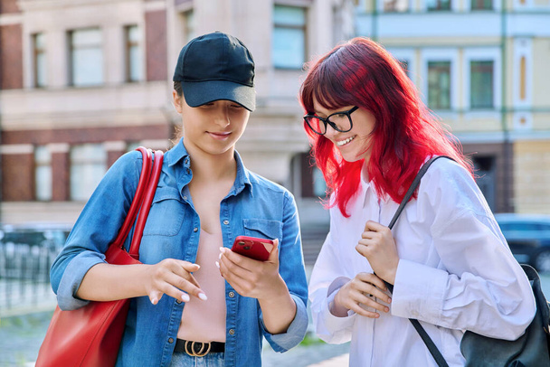 Two teenage females talking, looking into smartphone, outdoor on city street. Attractive girls friends, communicate together. Friendship, communication, adolescence, urban lifestyle, youth concept - Foto, imagen