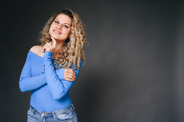 Woman in Blue Sweater Hugs Herself on Black Background. Smiling Happily Holding Hand to Face. Girl Smiles with Problematic Skin. Facial Skin Care. - Photo, image