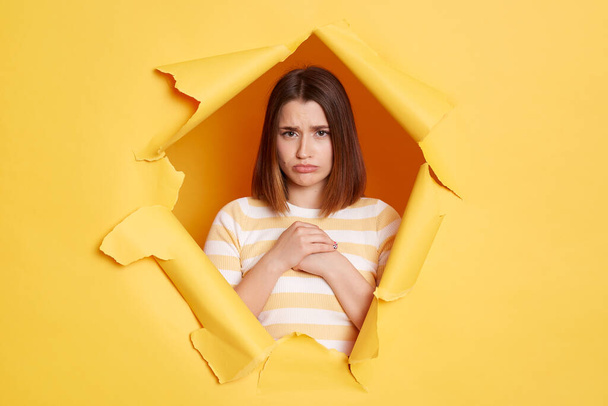 Portrait of sad upset woman wearing striped shirt posing in yellow paper hole, standing and and looking at camera with pout lips, expressing sorrow and sadness. - Foto, Bild