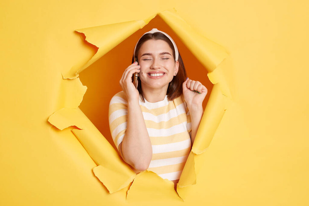 Indoor shot of smiling delighted satisfied woman wearing striped shirt and hair band posing in yellow paper hole, raised arm and talking on cell phone, being happy and glad to hear something pleasant. - Фото, зображення