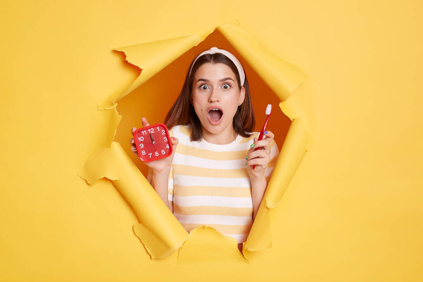 Portrait of shocked woman wearing striped shirt and hair band posing in yellow paper hole, holding alarm clock and toothbrush, looking at camera with open mouth, being hurry. - Foto, Imagen