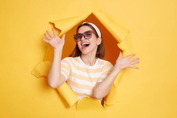 Portrait of attractive happy woman wearing striped shirt and hair band posing in yellow paper hole, standing with raised arms, celebrating her victory, expressing positive emotions. - Photo, image
