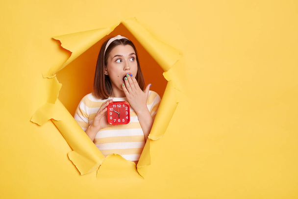 Indoor shtot of surprised woman wearing striped shirt and hair band posing in yellow paper hole, holding alarm clock in hand, covering mouth with palm, looking away at mockup for advertisement. - Foto, Bild