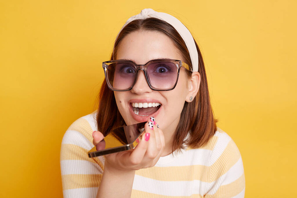 Portrait of happy smiling delighted woman wearing striped shirt posing isolated over yellow background, using cell phone for recording voice message, looking at camera with excited look. - Foto, Bild