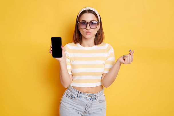 Romantic woman wearing striped shirt posing isolated over yellow background, showing smart phone with advertisement area on blank scree, sending air kisses, making love korean gesture. - Φωτογραφία, εικόνα