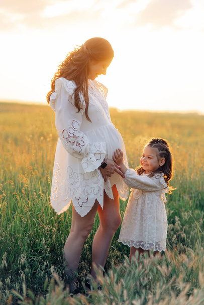 Little child girl touches belly of her pregnant mother among meadow at sunset. - 写真・画像