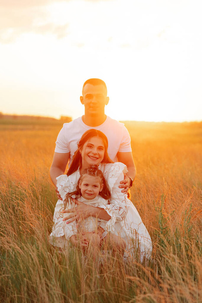 Joyeux jeune famille avec petite fille marche sur la prairie et s'assoit au milieu de l'herbe au coucher du soleil. - Photo, image