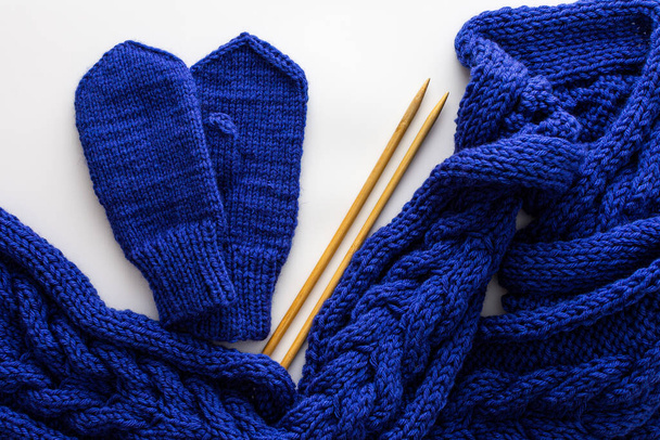 Blue knitted woolen scarf and mittens and knitting needles on a white background, homemade needlework concept, top view - Photo, Image