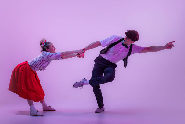 Gesellschaftliches Tanzen. Studioaufnahme eines jungen Mannes und einer jungen Frau in Vintage-Retro-Outfits, die auf fliederfarbenem Hintergrund in Neonlicht lindy hop tanzen. Zeitlose Traditionen, Mode und Kunst der 1960er Jahre - Foto, Bild