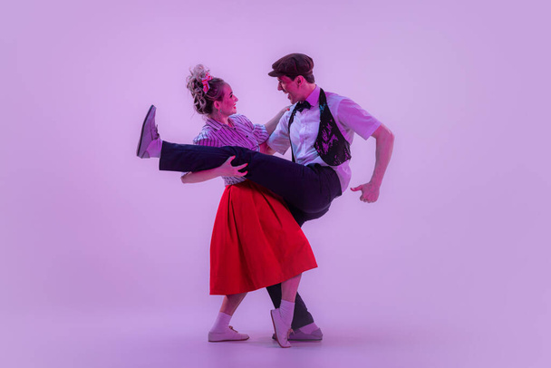 Energy, love. Stylish young man and pretty woman in vintage style outfits dancing retro dance isolated on very peri color background in neon light. Timeless traditions, 1960s fashion style and art - Foto, afbeelding