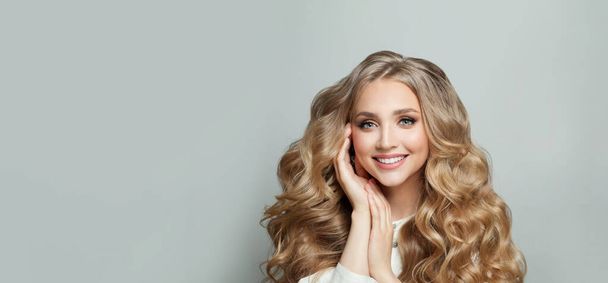 Closeup of happy pretty blonde young woman with long wavy hair smiling against white studio wall banner background - 写真・画像