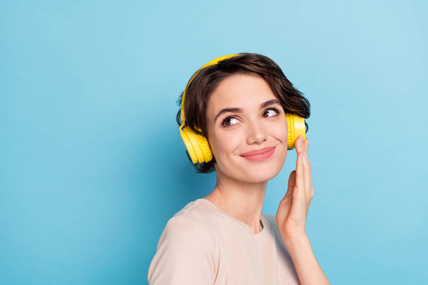 Photo of adorable dreamy lady dressed beige t-shirt earphones listening songs looking back empty space isolated blue color background. - Фото, зображення