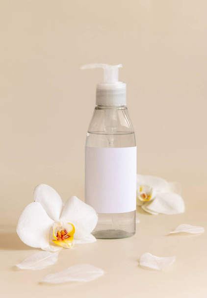 One pump dispenser bottle near white orchid flowers on light yellow close up, mockup. Skincare beauty product package. Exotic natural cosmetics, pastel minimal compositio - Фото, изображение