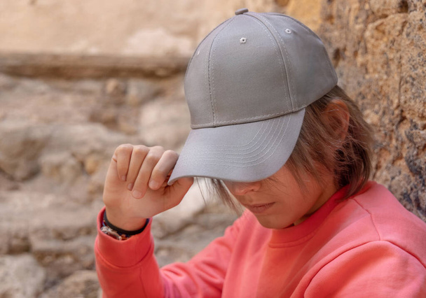Teen girl in grey baseball cap and pink long sleeve shirt looking into a camera. Cap mockup. Serious Teenager girl near a stone wall outdoor touching the visor. Loneliness concept - Foto, imagen