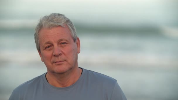 Close-up portrait of senior man against blurry ocean. Retired male with a faint smile touching his lips - Metraje, vídeo