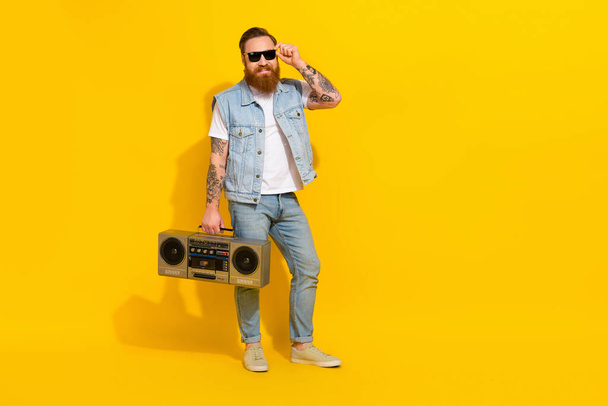 Photo of charming pretty man wear jeans waistcoat arm dark eyewear listening boom box empty space isolated yellow color background. - Foto, afbeelding