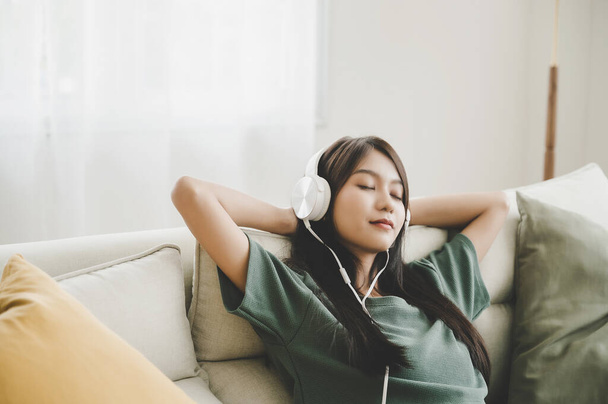 Mujer joven asiática sentada en un sofá. Feliz mujer escuchando música y durmiendo en la sala de estar. Ella feliz y relajante en el tiempo libre en fin de semana - Foto, Imagen