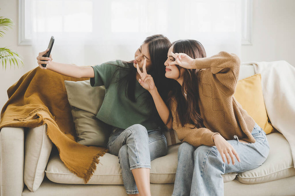 Дві азіатські молоді жінки щасливі посміхаються і приймають селфі на дивані у вітальні вдома
 - Фото, зображення