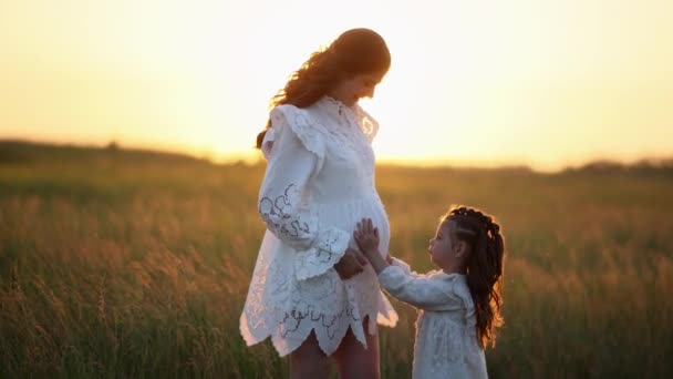 Petite fille enfant touche du ventre de sa mère enceinte et se blottit parmi la prairie au coucher du soleil. - Séquence, vidéo