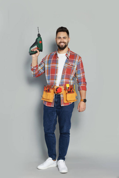 Young worker with power drill and tool belt on grey background - Φωτογραφία, εικόνα