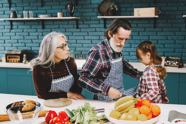 Mature couple and their granddaughter preparing food. Wearing aprons, modern kitchen interior. - Foto, immagini