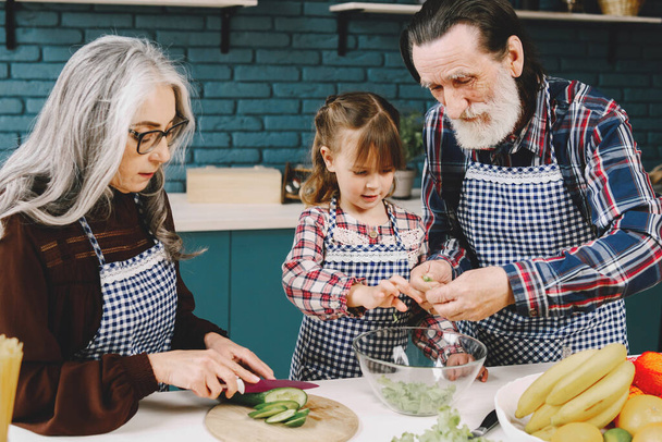 Mature couple and their granddaughter preparing food. Wearing aprons, modern kitchen interior. - Foto, Imagem