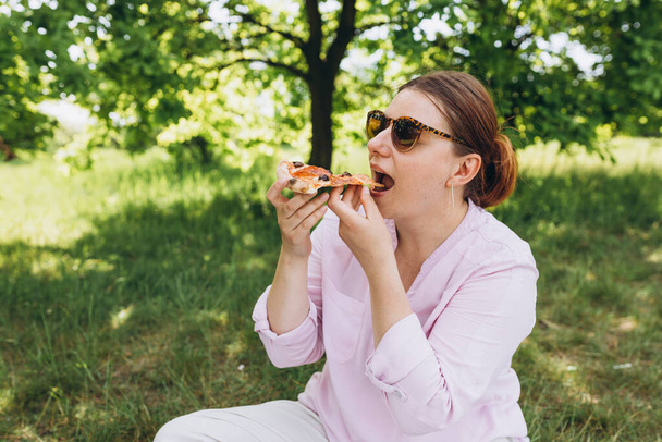 Chica morena divertida en gafas de sol comiendo pizza en el fondo de la naturaleza. Chica atractiva se siente hambriento y le encanta el sabor. Pizza entera para mí. Concepto de comida rápida - Foto, imagen