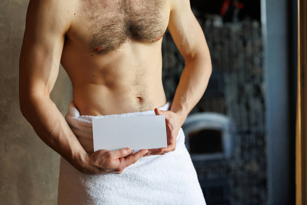 Uomo attraente con un busto nudo in un asciugamano bianco mostra un volantino vuoto - Foto, immagini